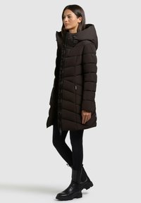 Cappotto lungo imbottito nero con collo alto e cappuccio, dotato di una zip frontale, cuciture diagonali e tasche laterali. Abbinato a pantaloni neri e stivali.
