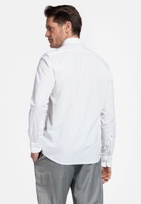 Witte blouse met lange mouwen, knoopmanchetten en een klassieke kraag. Zachte, ademende stof. Grijze broek. Achteraanzicht van de outfit.