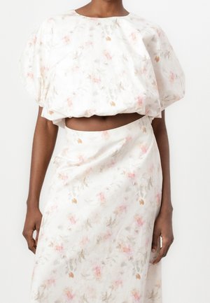 Femme portant un crop top blanc à manches bouffantes fleuri et une jupe taille haute assortie, montrant le ventre sur un fond uni.