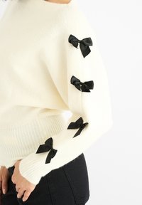 White sweater with a textured finish, featuring black satin bows on the sleeves. Knit cuffs and relaxed fit. Black jeans visible.