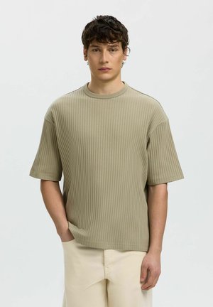 Jeune homme aux cheveux bouclés portant une chemise beige ample à côtes et manches courtes, ainsi qu’un pantalon clair, debout devant un fond clair uni.