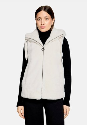 Femme portant une veste sans manches en fausse fourrure blanche zippée par-dessus un haut noir à manches longues et un pantalon noir, face à un fond uni.