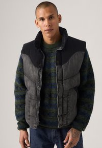 Chaleco acolchado de tela gris oscuro con cuello alto, cierre de cremallera y suéter verde con patrón debajo, con acentos en azul y tejido texturizado.