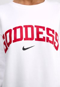 Valkoinen huppari, jossa on "GODDESS" paksuilla punaisilla kirjaimilla ja pieni musta Nike-logo. Pyöreä kaula-aukko ja pehmeä tekstuuri.