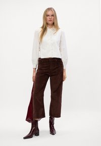 Witte textuurblouse met een kraag, bruine corduroy wijdbeenbroek en glanzende donkerbordeaux enkellaarsjes. Model staat met een tas.