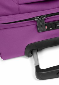 Purple rolling suitcase with a fabric exterior, dual zippers, and a retractable metal handle. Features a combination lock and textured accents.