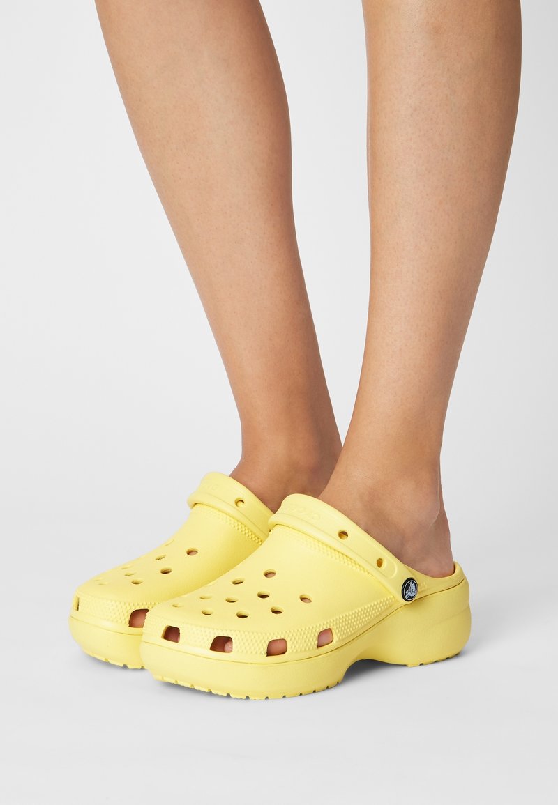 Jambes portant des sabots jaunes vifs à enfiler avec des trous de ventilation et une bride arrière, présentés sur un fond clair uni.