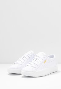 Weiße Sneaker mit einer glatten synthetischen Oberseite, Schnürdesign, strukturiertem Gummisohle und goldfarbenem Logo-Akzent an der Seite.