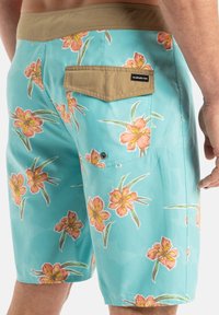 Krátke plavky v žiarivej tyrkysovej farbe s kvetinovým vzorom, ktorý obsahuje oranžové kvety. Obsahujú zadné vrecko so béžovým klopou.