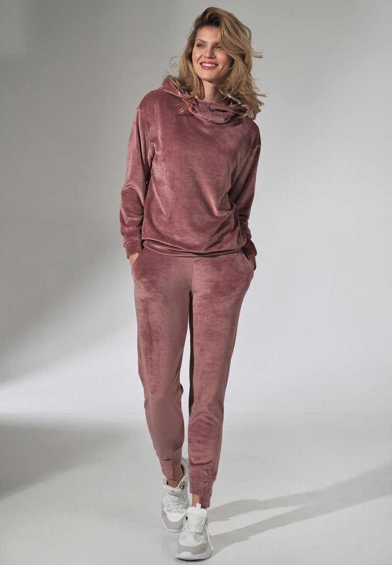 Welwetowy zestaw dresowy w kolorze przykurzonego różu, składający się z bluzki z kapturem i zwężanych spodni z kieszeniami, zestawiony z białymi trampkami. Gładka faktura.
