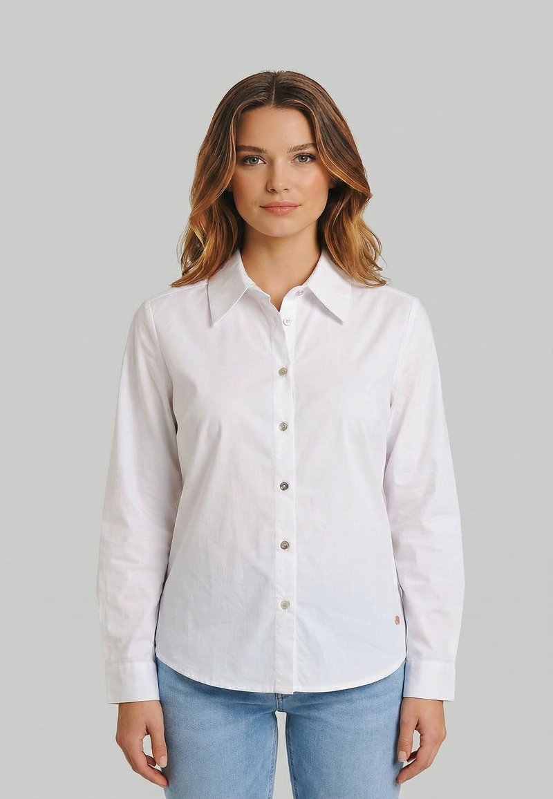 Donna con capelli castano chiaro che indossa una camicia bianca con bottoni e jeans azzurri, in piedi davanti a uno sfondo grigio uniforme.