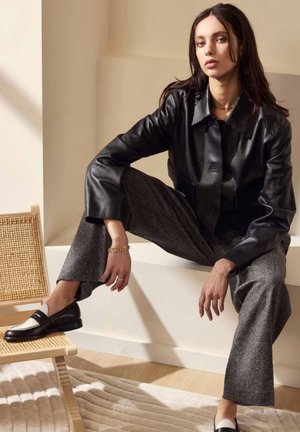 Veste courte en cuir noir, pantalons gris à jambes larges, mocassins noir et blanc. Modèle avec de longs cheveux, accessoires discrets, assis sur une chaise.