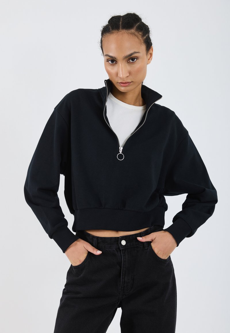 Sweatshirt court noir avec col montant et fermeture éclair argentée, associé à un jean noir taille haute, comportant des poches latérales et une fermeture à bouton.
