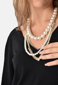Collane a tre strati con grandi perle bianche, alternate a catene color oro e piccoli accenti di cristallo, indossate su un top nero.