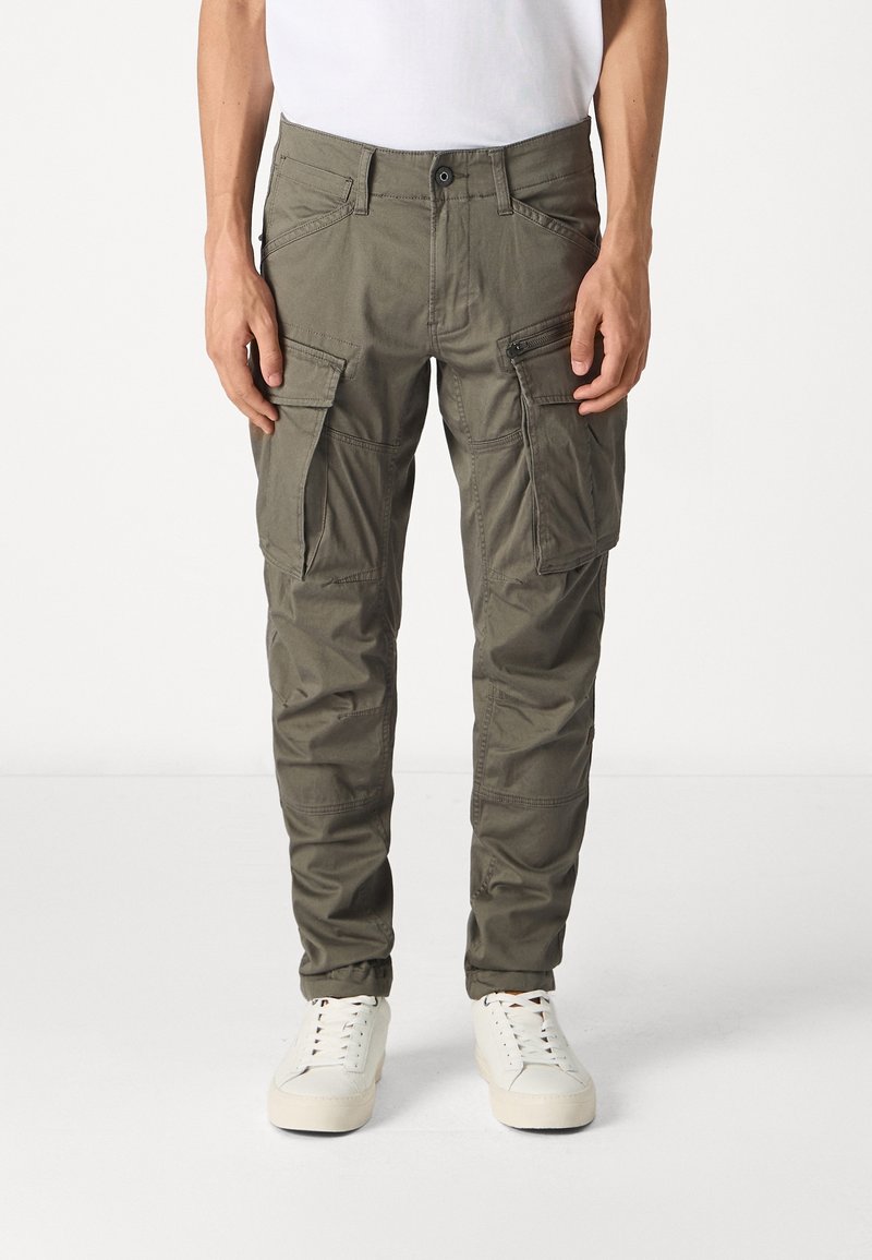 Personne portant un pantalon cargo vert olive avec plusieurs poches et des baskets blanches, debout devant un fond clair uni.