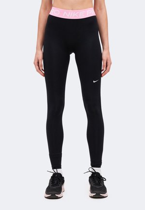Femme portant des leggings Nike noirs avec une ceinture rose et des chaussures de running noires, debout devant un fond uni.
