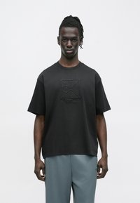 Μαύρο βαμβακερό μπλουζάκι με ανάγλυφο λογότυπο Calvin Klein στο κέντρο. Κοντά μανίκια, κλασικό στρογγυλό λαιμό, άνετη εφαρμογή. Ίσια γραμμή στο τελείωμα.