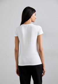 T-shirt bianco a maniche corte con scollo rotondo, silhouette aderente e texture liscia. Dettaglio del logo sull'orlo inferiore.