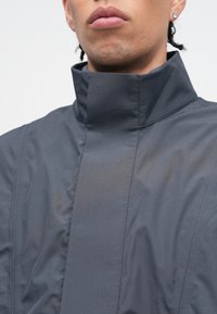 Tmavosivá bunda s vysokým golierom vyrobená z ľahkého materiálu, so čistými líniami, textúrovaným povrchom a minimalistickým dizajnom.