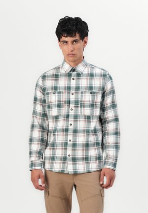 Homme portant une chemise blanche à manches longues avec un motif en tartan vert et rouge et un pantalon cargo beige, se tenant contre un fond blanc.