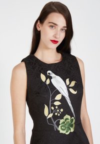 Vestido negro sin mangas con bordado floral texturizado, que presenta un pájaro blanco, hojas amarillas y acentos verdes en la parte delantera.