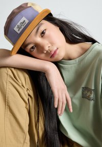 Casquette multicolore à visière plate avec une étiquette. Le modèle porte une chemise vert d'eau avec un design brodé et un pantalon beige.