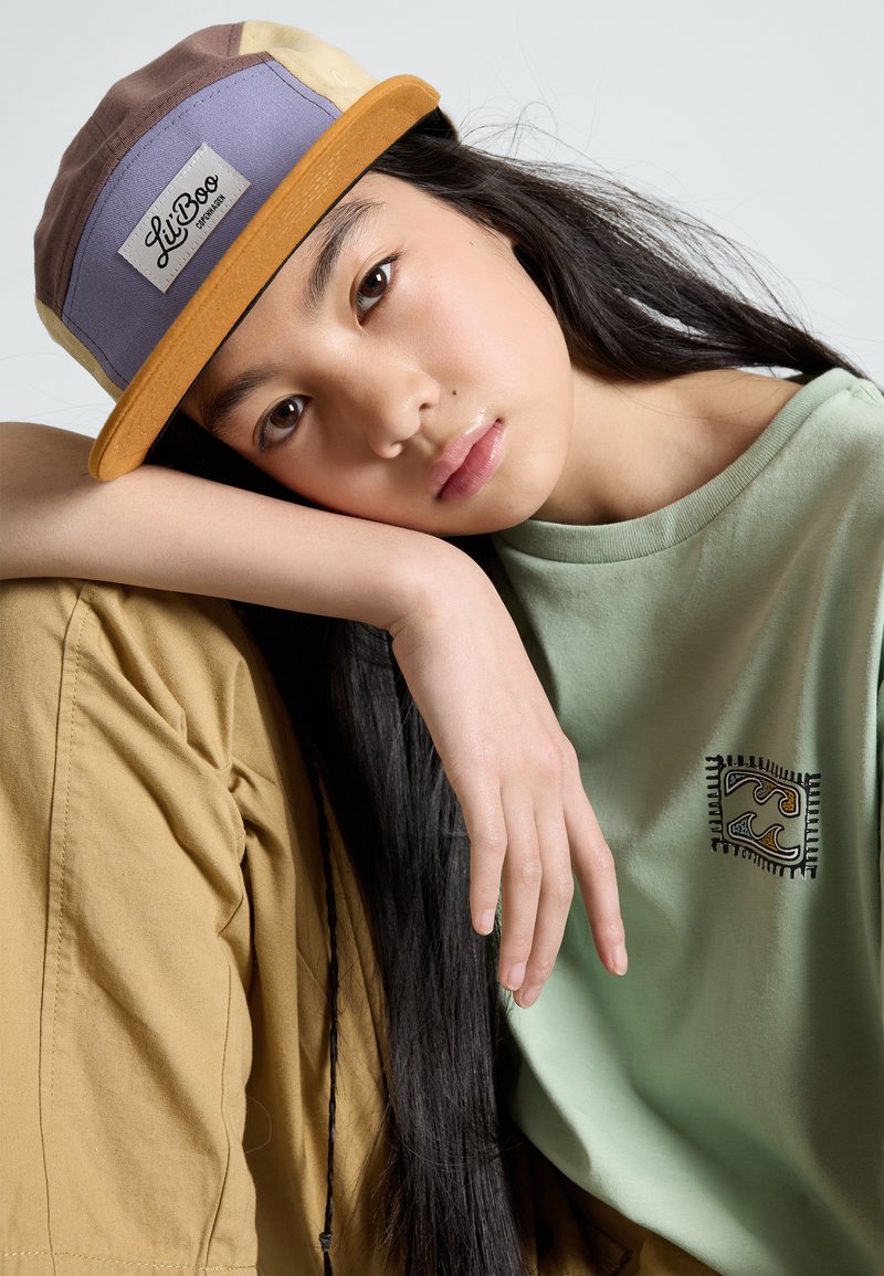 Casquette multicolore à visière plate avec une étiquette. Le modèle porte une chemise vert d'eau avec un design brodé et un pantalon beige.