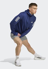 Sportska plava polu-zip dukserica s bijelim trostrukim trakama. Lagan, glatki materijal. Uparena s ljubičastim šortsevima i bijelim sportskim cipelama.