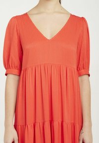 Červené plisované šaty s výstřihem do V a balónovými rukávy, které mají volnou vrstvenou sukni a lesklý povrch.