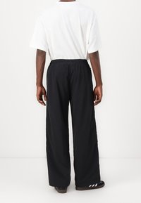 Svarta vida byxor i lätt material, med resår i midjan. Bärs tillsammans med en enkel vit skjorta och bruna slidesandaler.