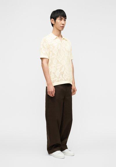 Homme debout portant un polo en maille crème à motif floral, un pantalon ample marron foncé et des baskets blanches, sur un fond clair uni.