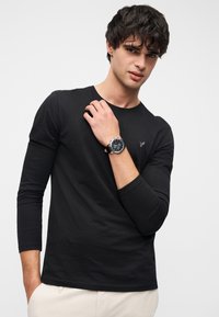 Tricou negru cu mâneci lungi, realizat dintr-un material moale, cu un mic logo pe piept. Modelul poartă un ceas de mână cu cadran negru și curea de piele.