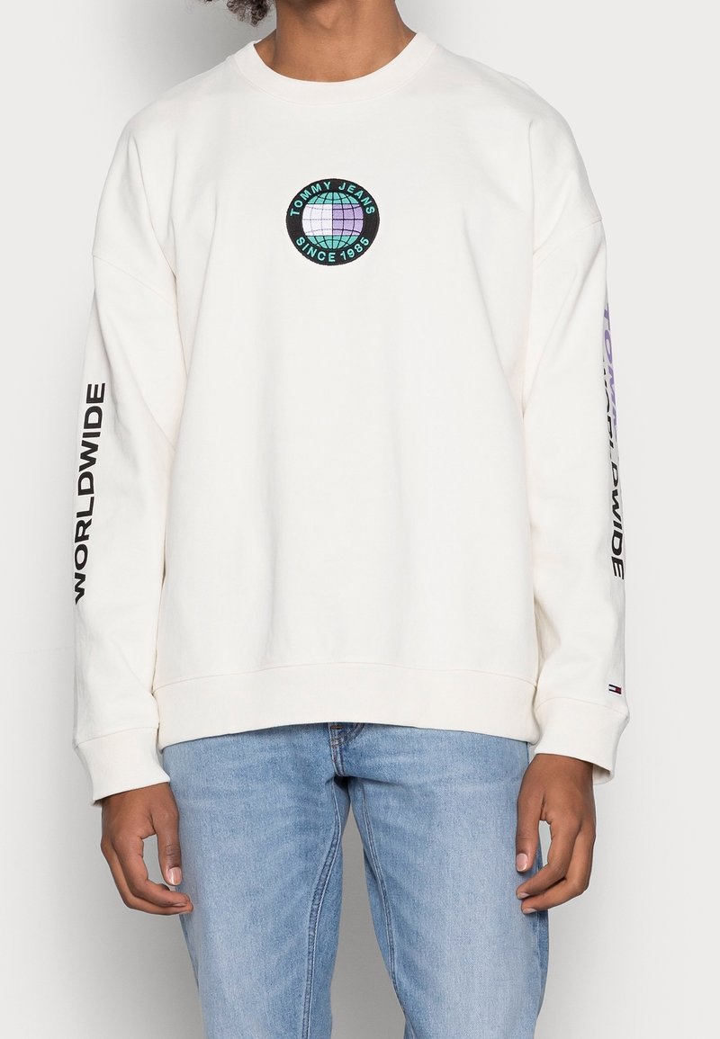 Felpa crema con un logo circolare multicolore sul petto. Le maniche lunghe presentano la scritta "WORLDWIDE" in nero. Il tessuto sembra morbido; design casual.