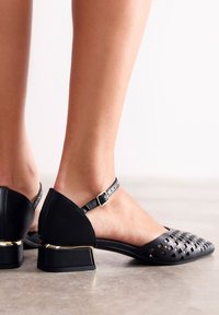 Zapatos negros con correa al tobillo, diseño recortado, tacón bajo en bloque y un acento dorado brillante en el tacón. Textura suave, forma elegante.