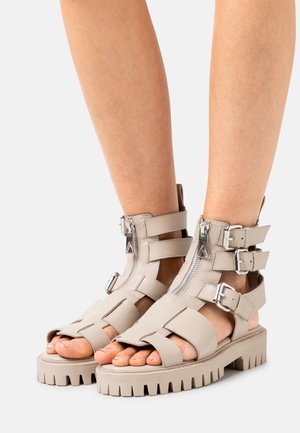 Vaaleanbeige nahkasaappaat, joissa on vetoketj closures, useita nauhaaksentteja, avonainen varvaskuvio ja paksu, uurrettu pohja.