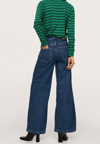 Osoba v zeleno-černém pruhovaném tričku s dlouhým rukávem, s vysokým pasem a širokými modrými džíny, a černých kožených kotníkových botách na podpatku, stojí s jednou rukou v kapsách.