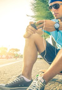 Young man wearing a blue shirt and sunglasses, sitting on a skateboard, holding a device with earbuds. Gray sneakers and short dark shorts.