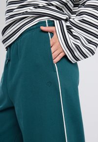 Hand in pocket of teal sweatpants with white side stripe, paired with black and white striped long-sleeve shirt.