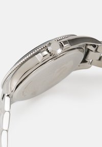Reloj de acero inoxidable plateado con caja pulida, bisel texturizado y corona plana, que cuenta con una parte trasera circular lisa y una pulsera de eslabones.