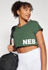 Žalias trumpas T-shirt su baltu "NEB" tekstu, trumpomis rankovėmis ir nelygiu apvadu. Modelis laiko juodą sportinį krepšį. Balti aukšto juosmens legginai.