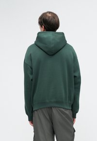Sweatshirt verde com uma grande capuz, ajuste descontraído e cintura canelada. Usado com calças cargo cinza claro. A textura parece suave e casual.