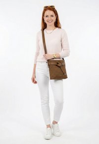 Kvinde med rødt hår iført lyserød trøje, hvide bukser, hvide sneakers, solbriller på hovedet og bærende en brun crossbody taske, smilende.