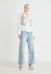 Camicia bianca a maniche lunghe con colletto a volant, gilet corto argentato, jeans a gamba larga azzurri e sandali con plateau argentati.