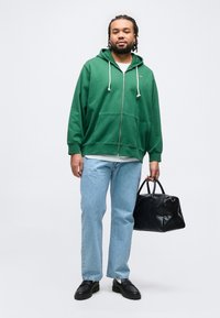 Άνδρας με γενειάδα και πλεγμένα μαλλιά, ντυμένος με πράσινο φούτερ με φερμουάρ και ανοιχτόχρωμο τζιν, κρατώντας μαύρη υφασμάτινη τσάντα, σε απλό φόντο.