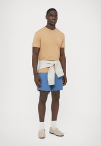 Tanfarget bomulls-t-skjorte med liten logo, grå hettegenser knyttet rundt livet, blå shorts og beige loafers med hvite sokker.