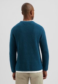 Man in een gestructureerde donkerteal trui over een wit shirt en beige broek, met zijn rug naar de camera gekeerd tegen een effen lichtgrijze achtergrond.
