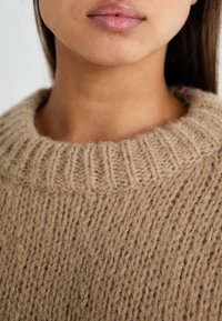 Chunky strikkegenser i lys beige, med ribbestrikket rund hals og et strukturert vevd mønster. Myk, varm ullstoff.
