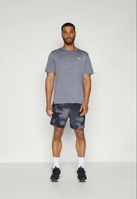 Hombre de pie, vistiendo una camiseta gris de Under Armour, pantalones cortos de camuflaje negro y gris, calcetines blancos y zapatillas de correr negras frente a un fondo plano.