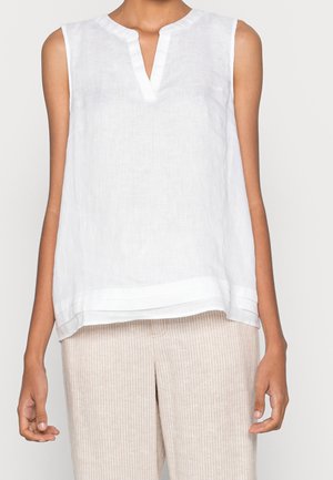 Mouwloze witte linnen blouse met ingesneden halslijn gedragen met lichtbeige gestreepte broek bij een persoon.