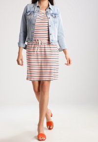 Robe rayée en tissu léger, avec des rayures rouges et marine, dotée d'une taille à cordon. Portée avec une veste en denim claire et des sandales oranges.